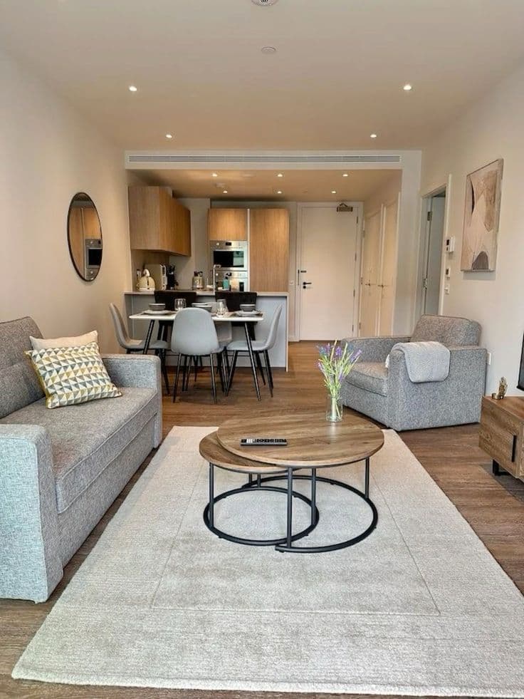 Séjour d'un appartement de centre-ville : canapé et fauteuil gris autour d'une table basse ronde, coin repas et cuisine ouverte au fond